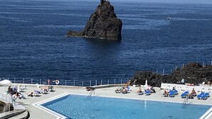 A pool area with clear blue water, surrounded by lounge chairs and umbrellas, with a rocky coastline in the background. - Penthouse Caniço de Baixo (Santa Cruz)