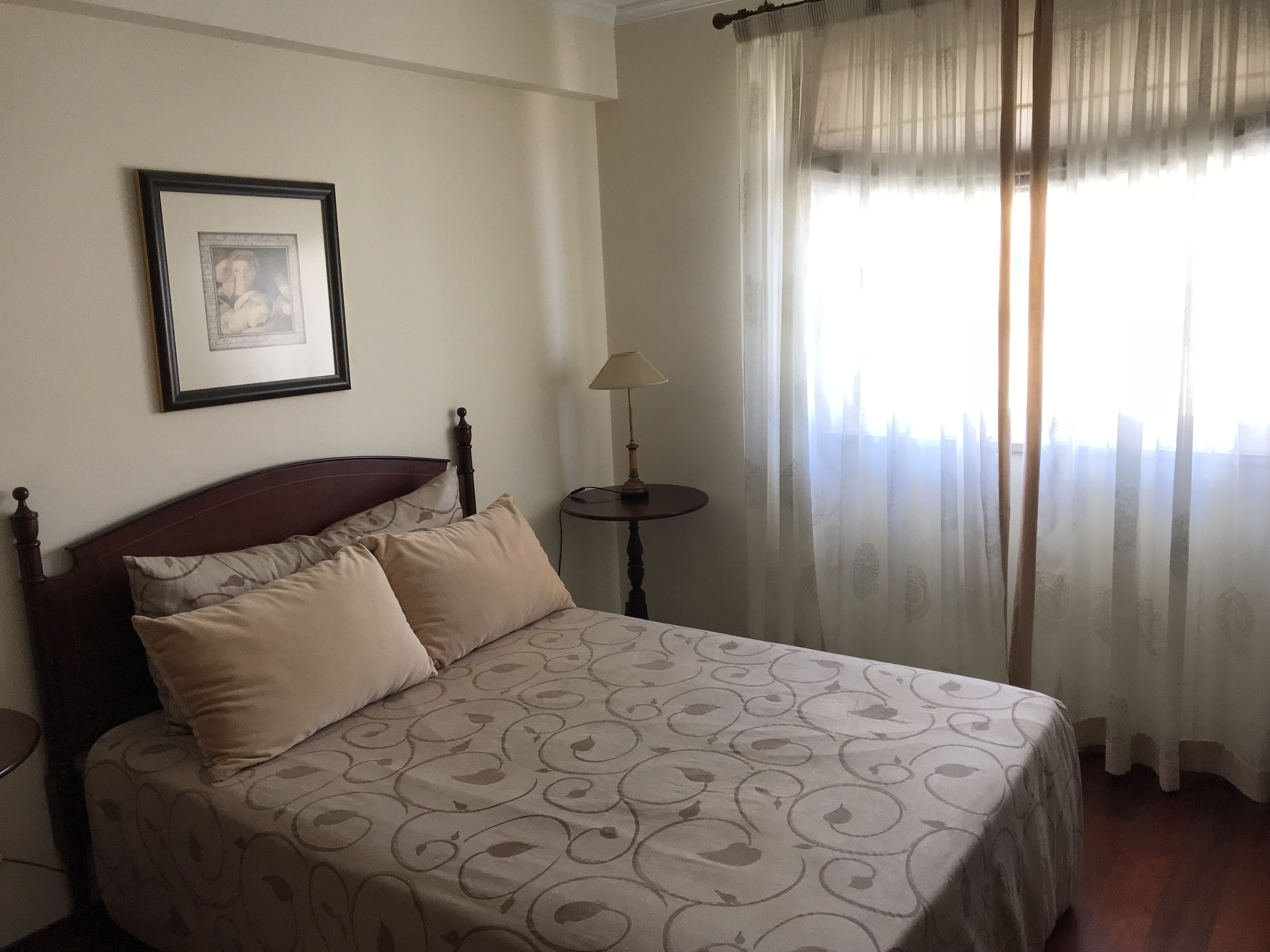 Un dormitorio con cama, una mesita de noche, lámpara, un cuadro enmarcado en la pared y cortinas traslúcidas en la ventana.