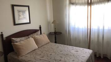 Un dormitorio con cama, una mesita de noche, lámpara, un cuadro enmarcado en la pared y cortinas traslúcidas en la ventana.