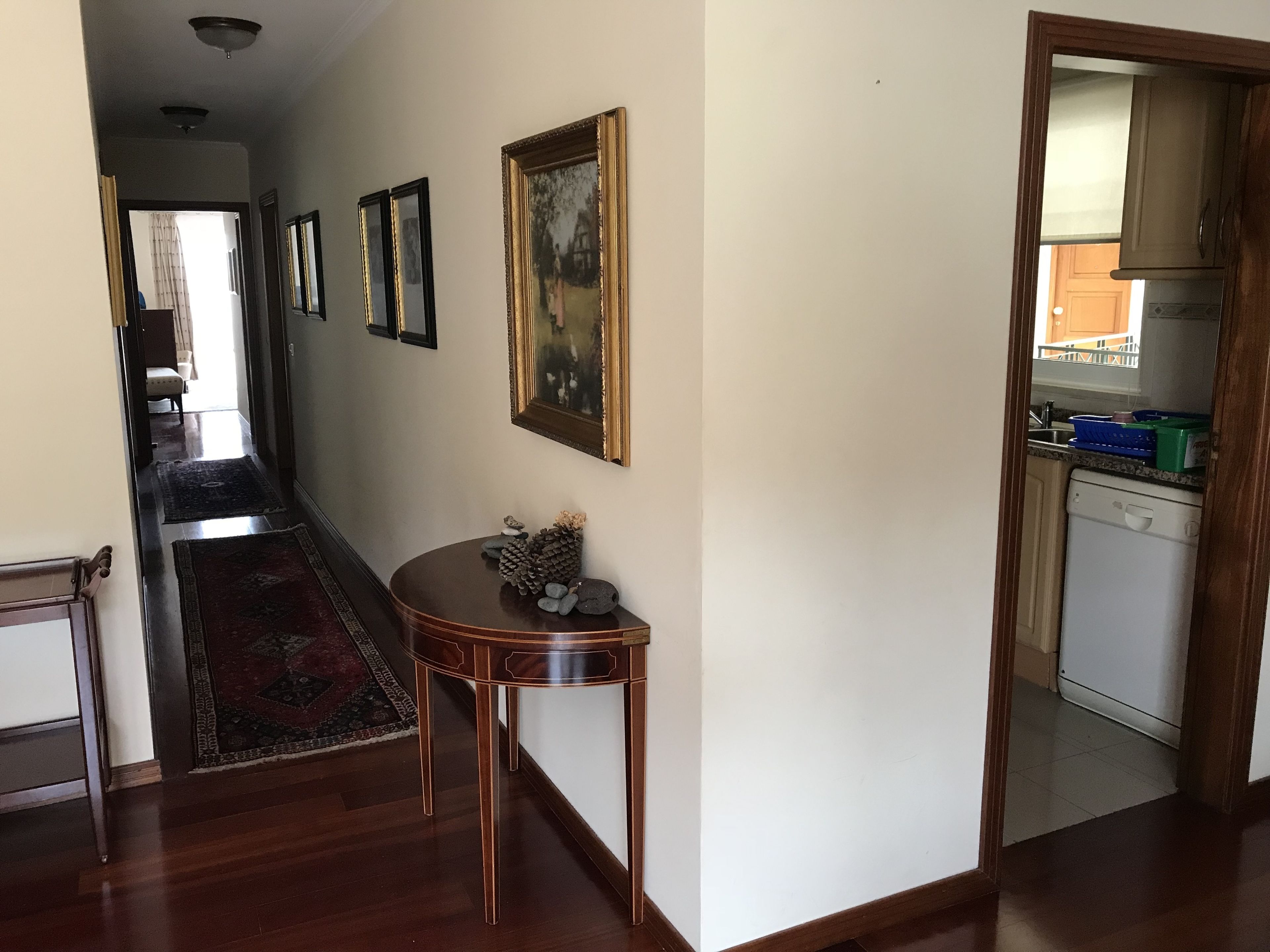 A hallway with a wooden floor, a rug, a small table with decorative items, and framed pictures on the wall.