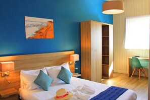 A neatly made bed with pillows, a hat, and towels, a wooden wardrobe, a desk with a chair, and a painting on the wall.