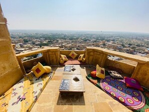 A terrace with a view of a cityscape, featuring a low table, cushions, and a patterned rug. - Desert Haveli (Jaisalmer)