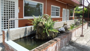 A building with a brick exterior, a window with a metal grill, and a small water feature with rocks and plants. - Dulan Red House (Donghe)