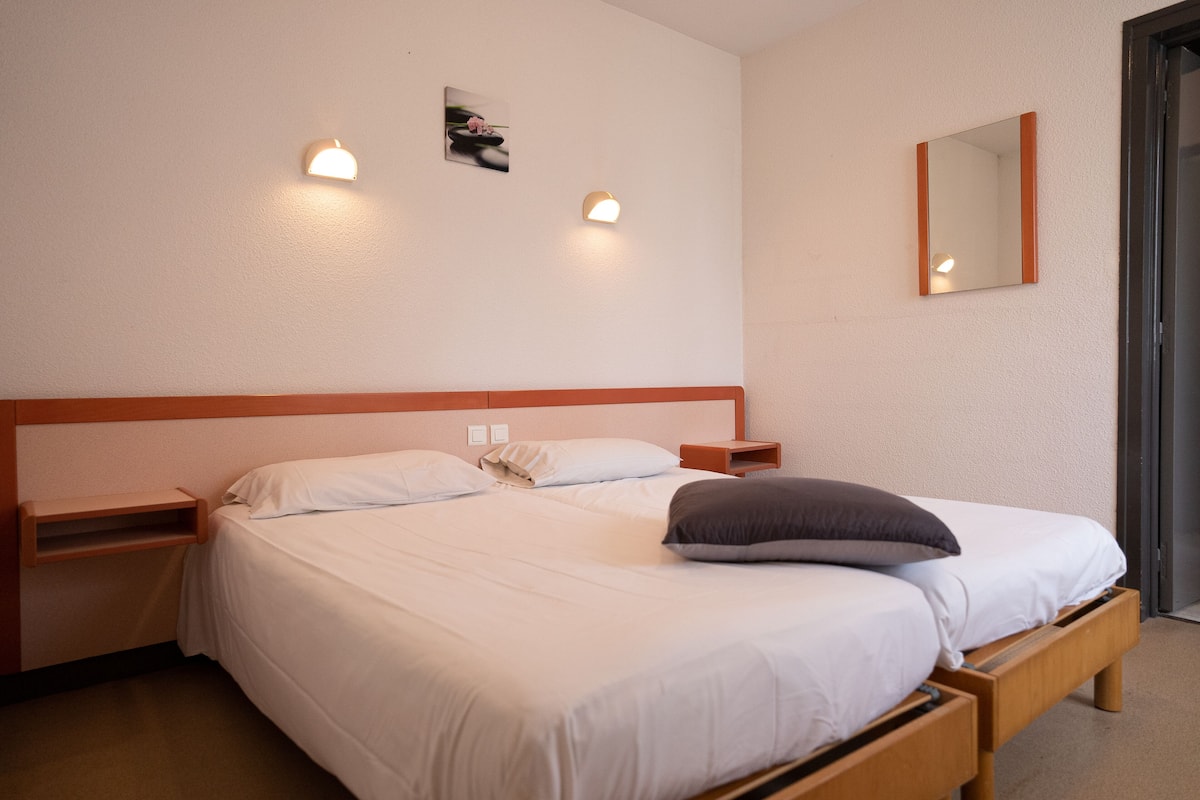 Un lit bien fait, avec du linge de lit blanc et une tête de lit en bois, deux tables de chevet avec des lampes, un petit tableau accroché au mur et un miroir.