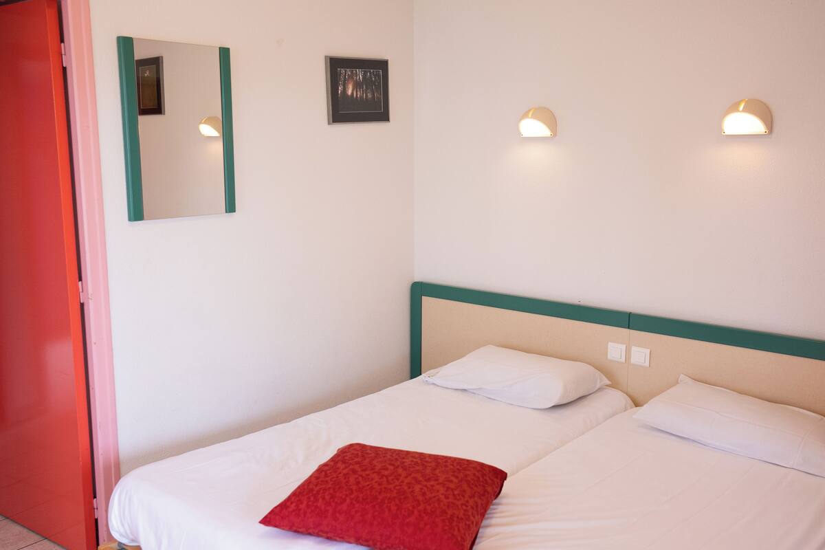 Une chambre d’hôtel avec deux lits, un oreiller rouge, un miroir et des lampes fixées au mur.