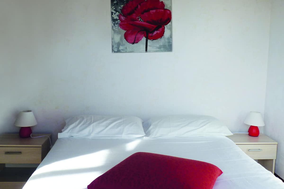 Une chambre à coucher avec un lit, des oreillers blancs, un plaid rouge, deux tables de chevet avec des lampes et un tableau représentant une fleur rouge accroché au mur.