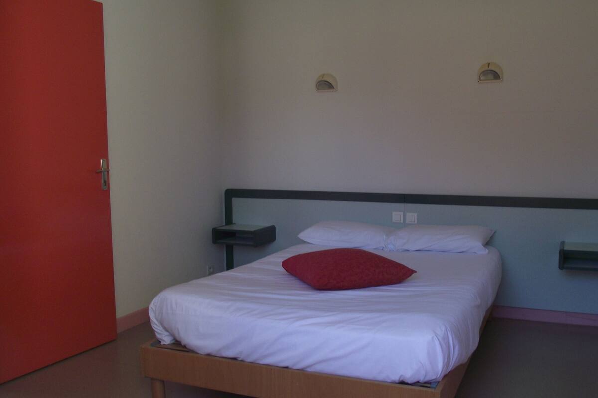 Un lit simple avec un cadre en bois, une literie blanche et un oreiller rouge. Il y a deux tables de chevet avec des lampes. Une porte rouge est visible sur la gauche.