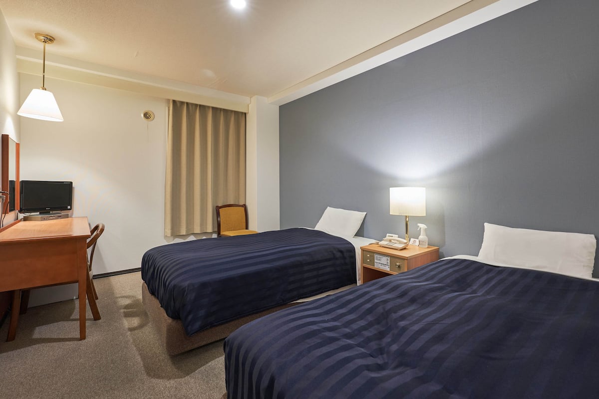 A hotel room with two beds, a desk with a computer, a chair, a lamp, and a nightstand with a phone and a bottle.
