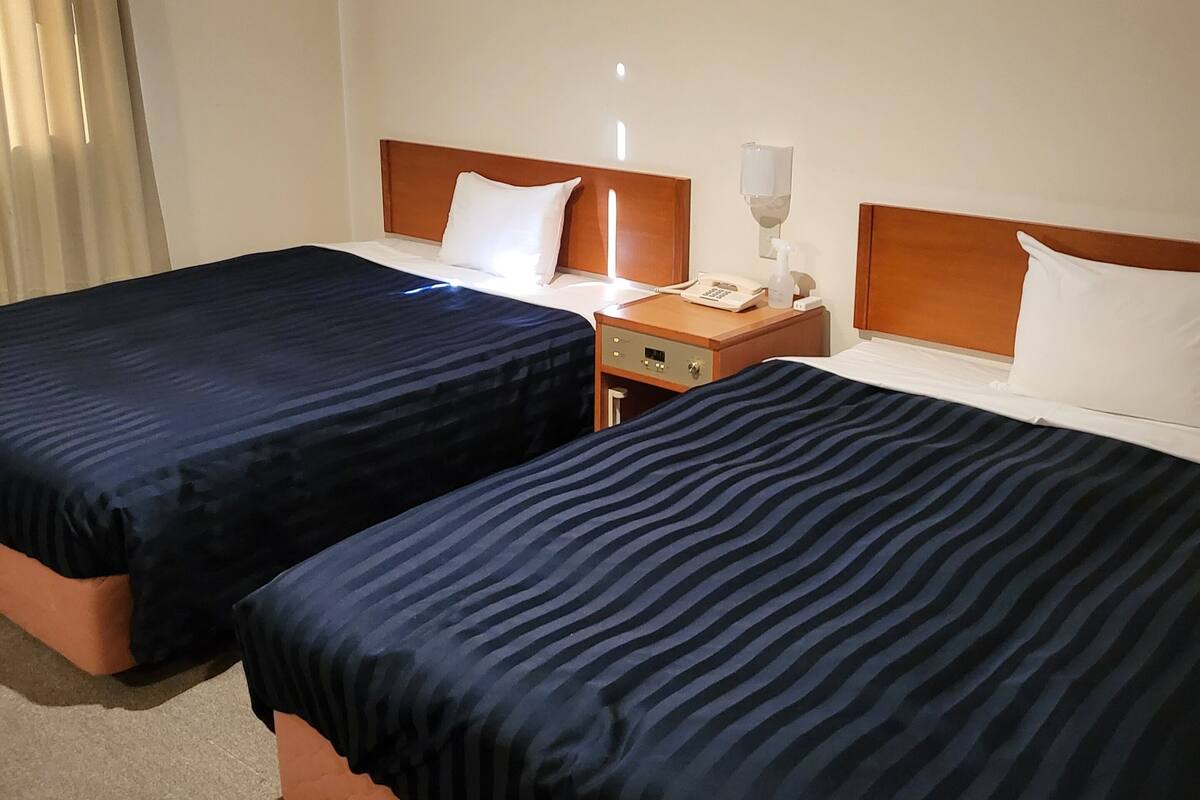 A hotel room with two beds, a nightstand, a lamp, and a window with curtains.