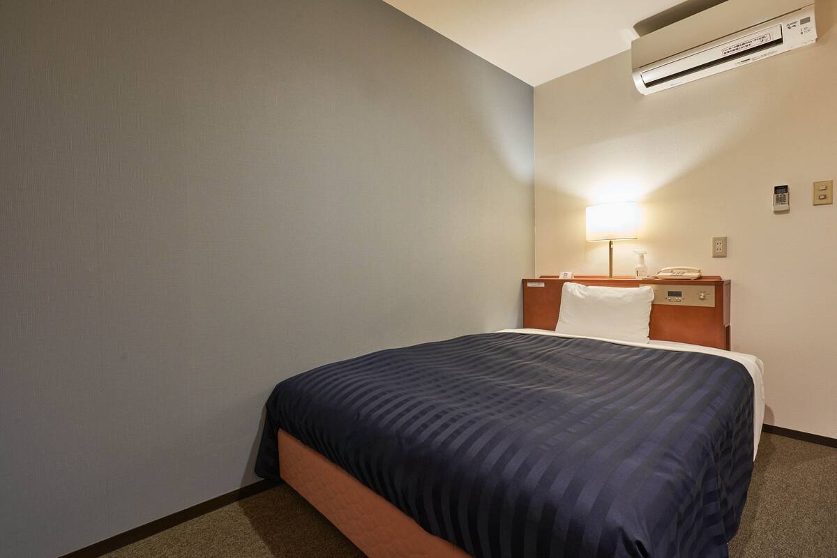 A hotel room with a bed, a nightstand, a lamp, and an air conditioning unit.