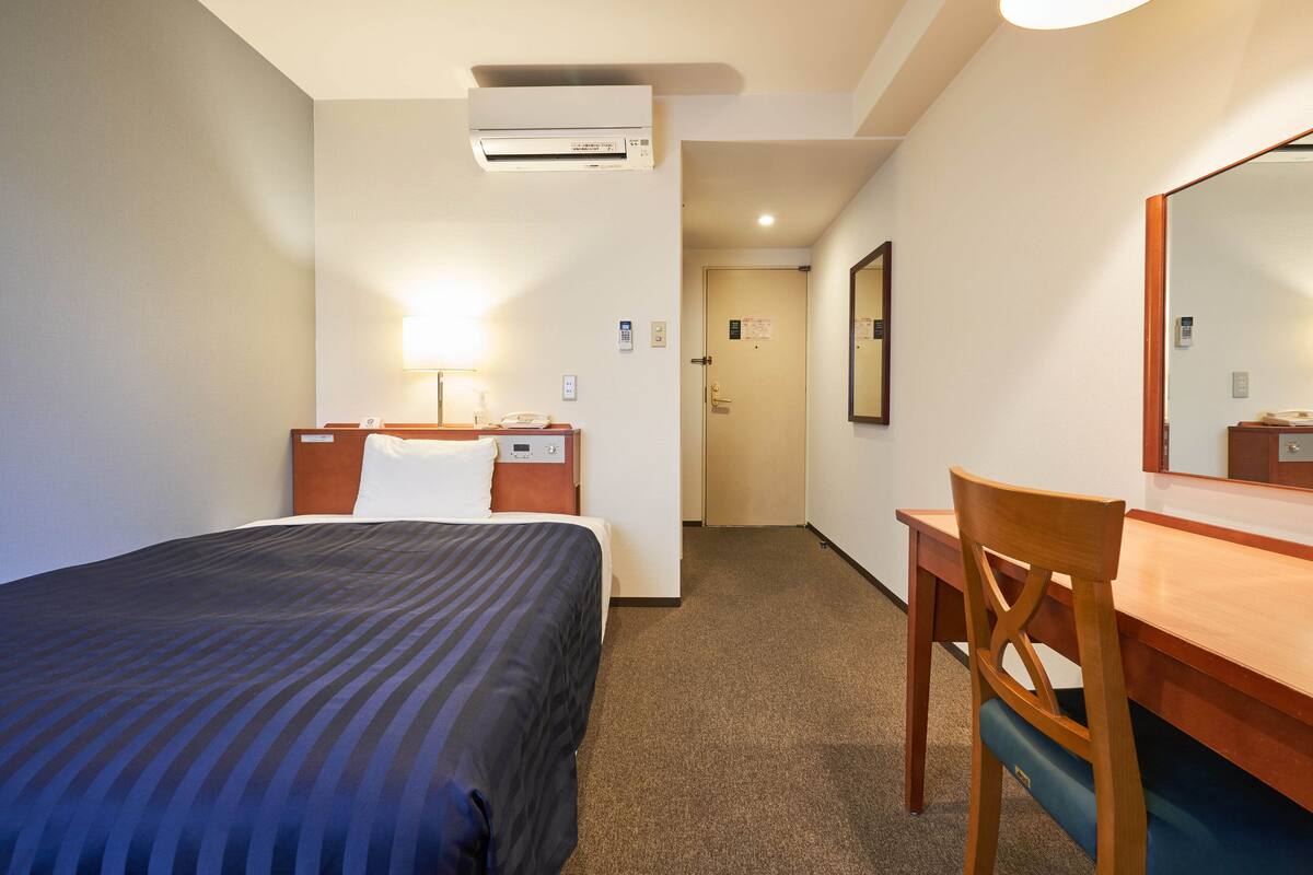 A hotel room with a bed, a desk, a chair, a mirror, and an air conditioning unit.