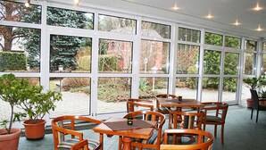 A covered outdoor seating area with tables and chairs, large windows, and potted plants. - Hotel Rodewisch (Rodewisch)
