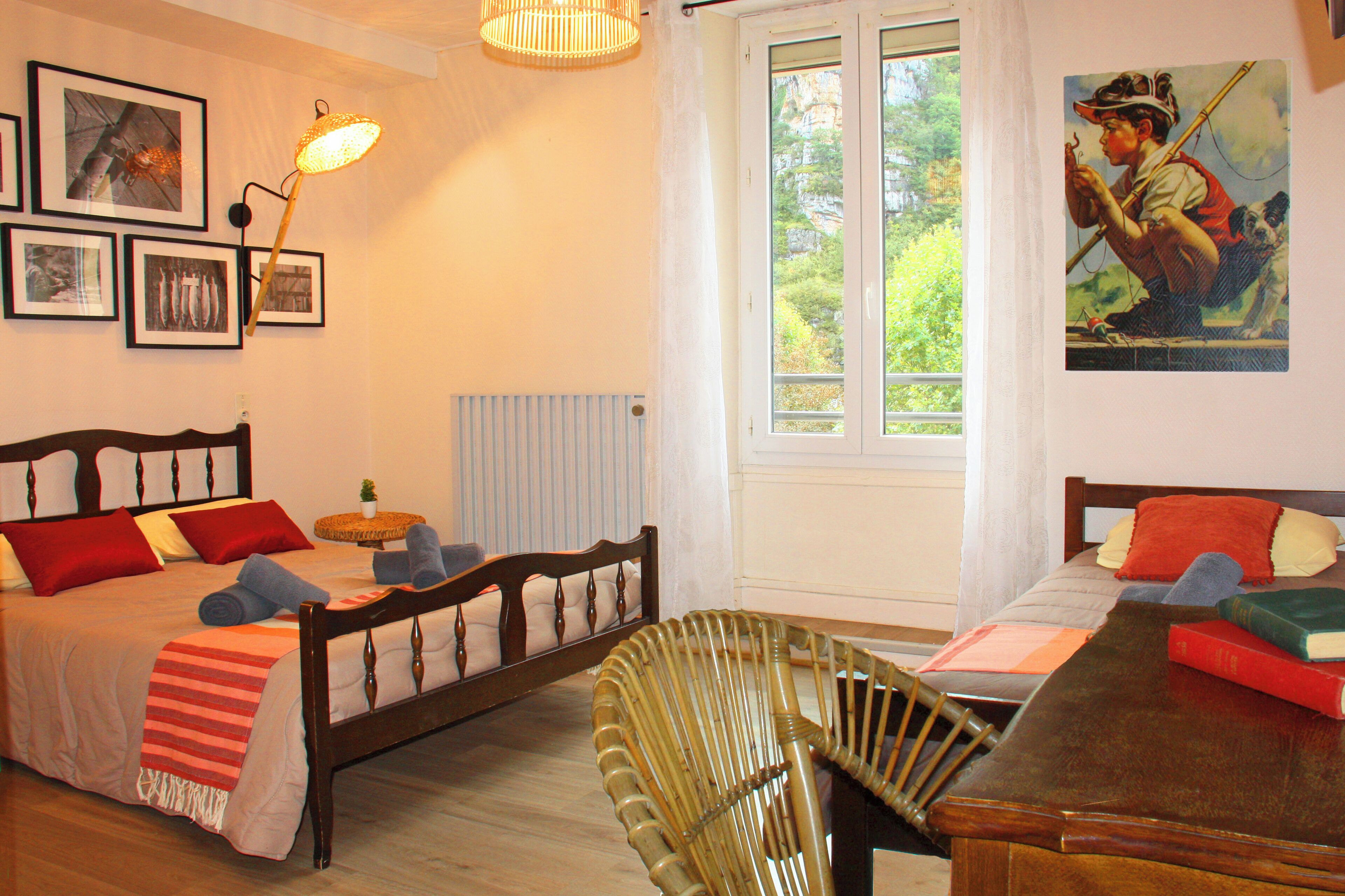 A bedroom with a bed, a chair, a table, a lamp, and a window with a view of greenery.
