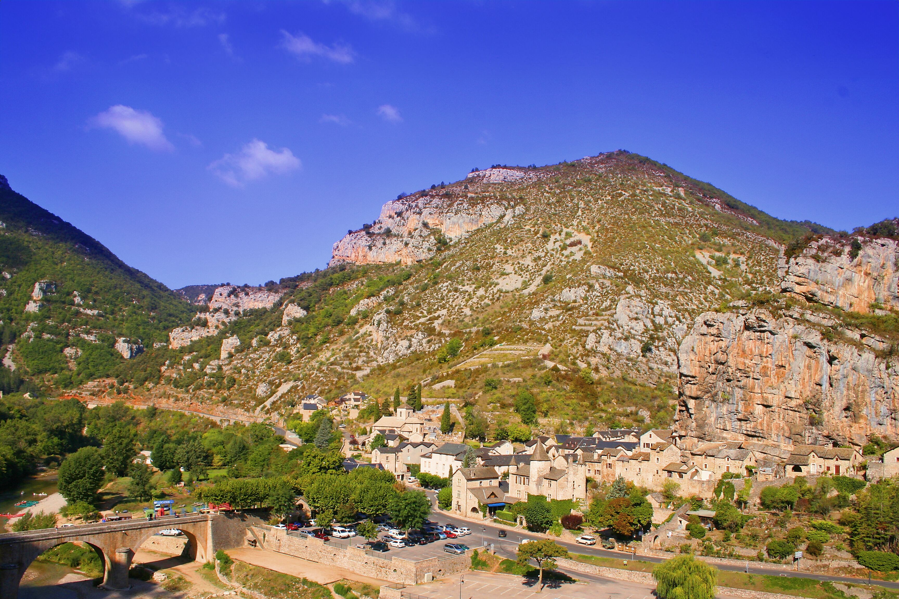 Une ville nichée dans une vallée, dominée par une montagne imposante, où l’on trouve un pont, un parking et un mélange de bâtiments résidentiels.