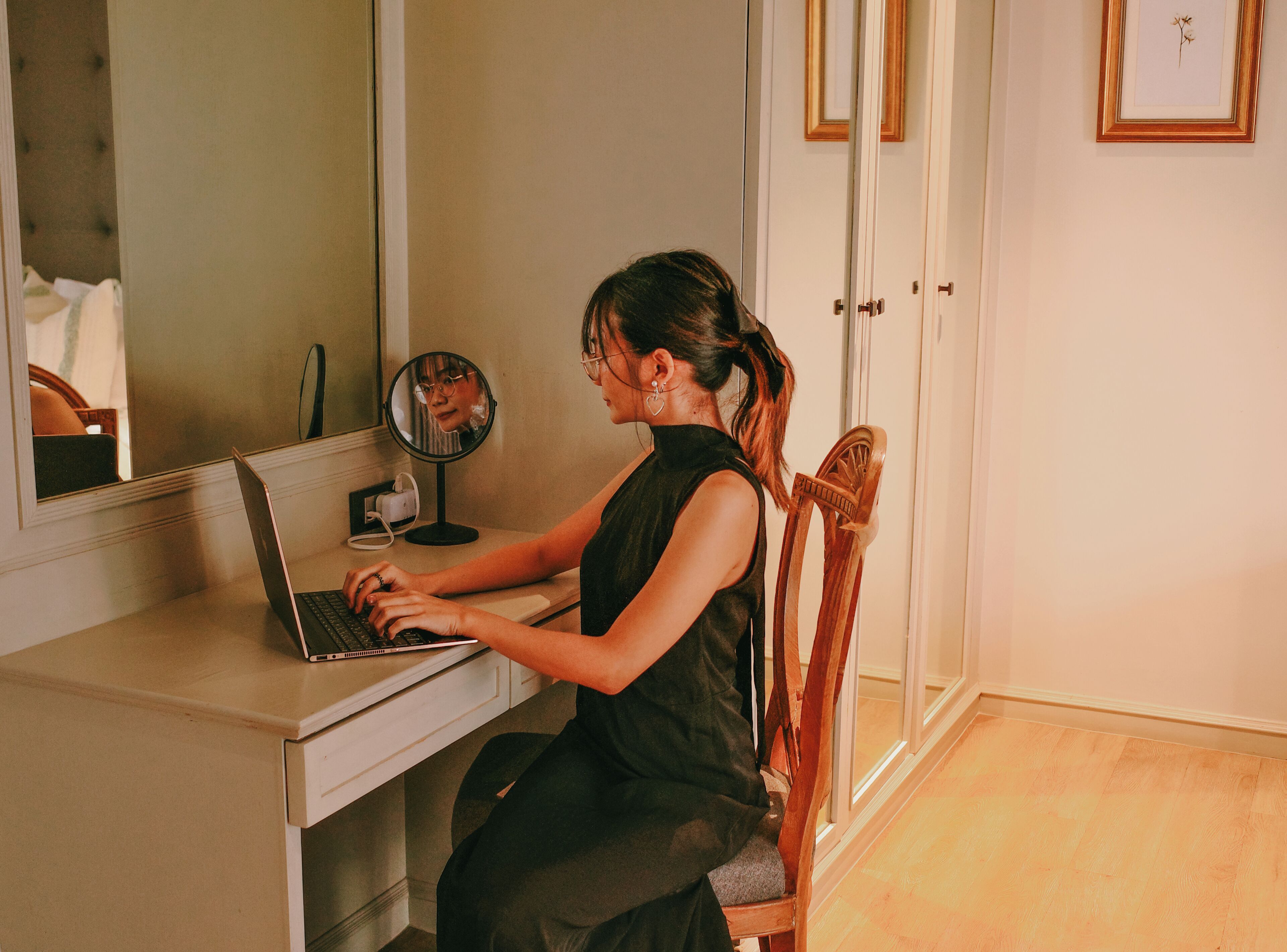 Uma pessoa sentada em uma mesa com um laptop, um espelho e uma pequena mesa com uma xícara.