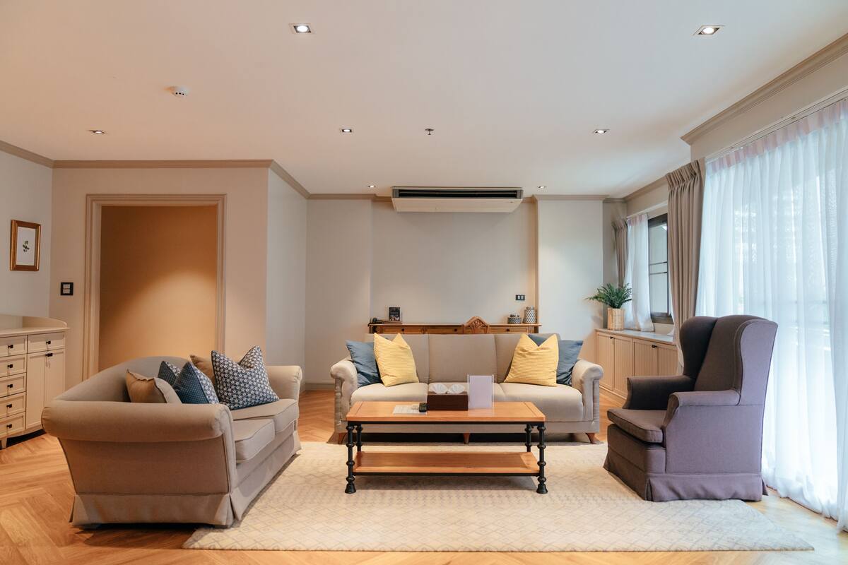 Ein modernes Wohnzimmer mit einem beigefarbenen Sofa, einem passenden Sessel und einem Couchtisch aus Holz. Ein großes Fenster mit durchsichtigen Vorhängen, eine integrierte Klimaanlage und eine Holzkommode mit Dekorationselementen.