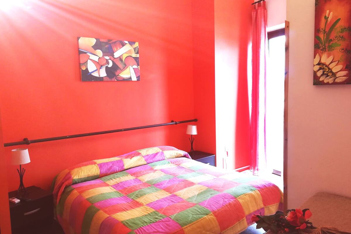 Una habitación con cama, mesita de noche, lámpara, un cuadro y una ventana con cortinas.