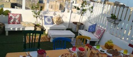 Una terraza en la azotea con una mesa lista para una comida, rodeada de vallas blancas y plantas en macetas.