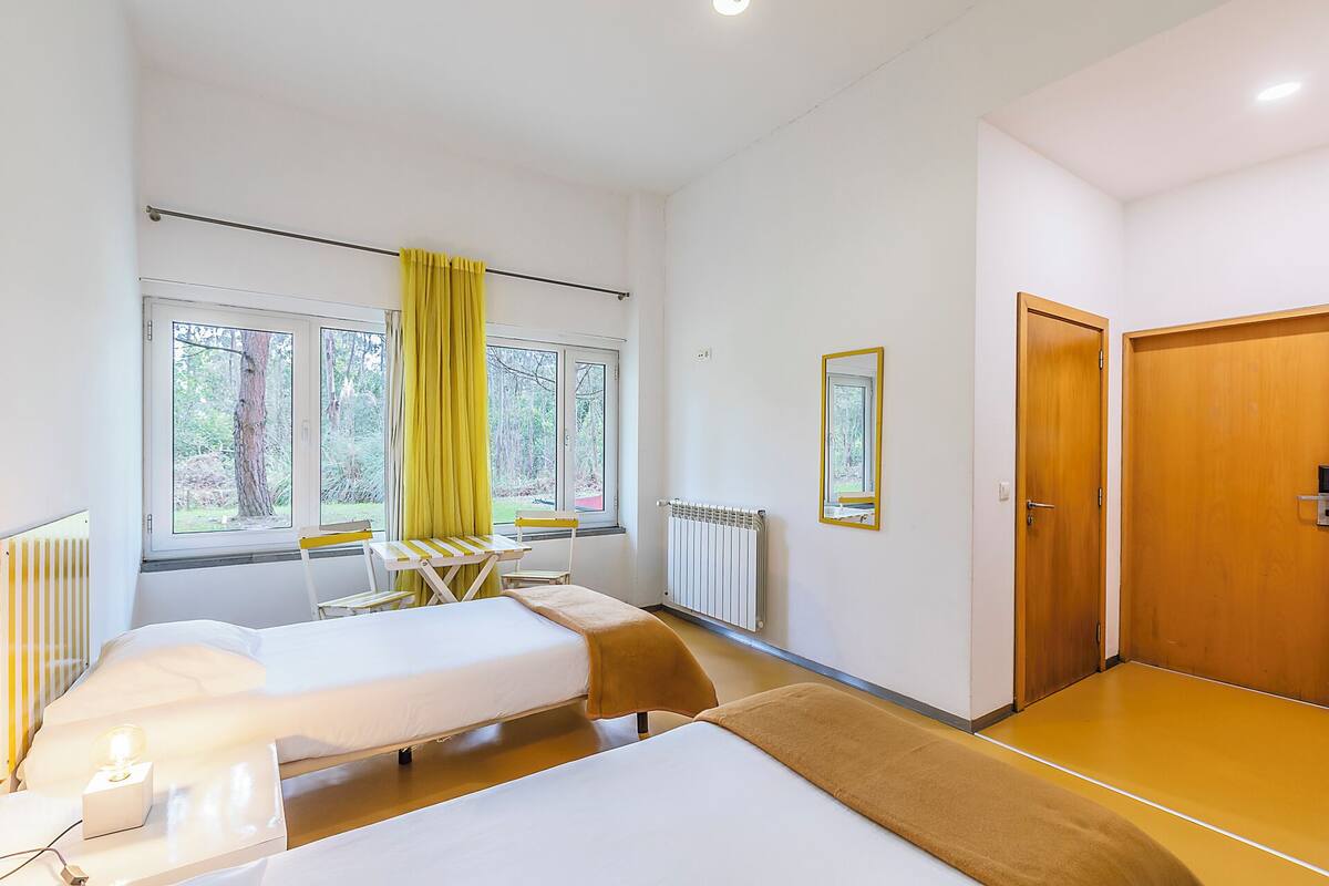 Ein Hotelzimmer mit zwei Betten, einem Tisch, Stühlen, einem fenster mit Vorhängen, einer Heizung und einer Holztür.