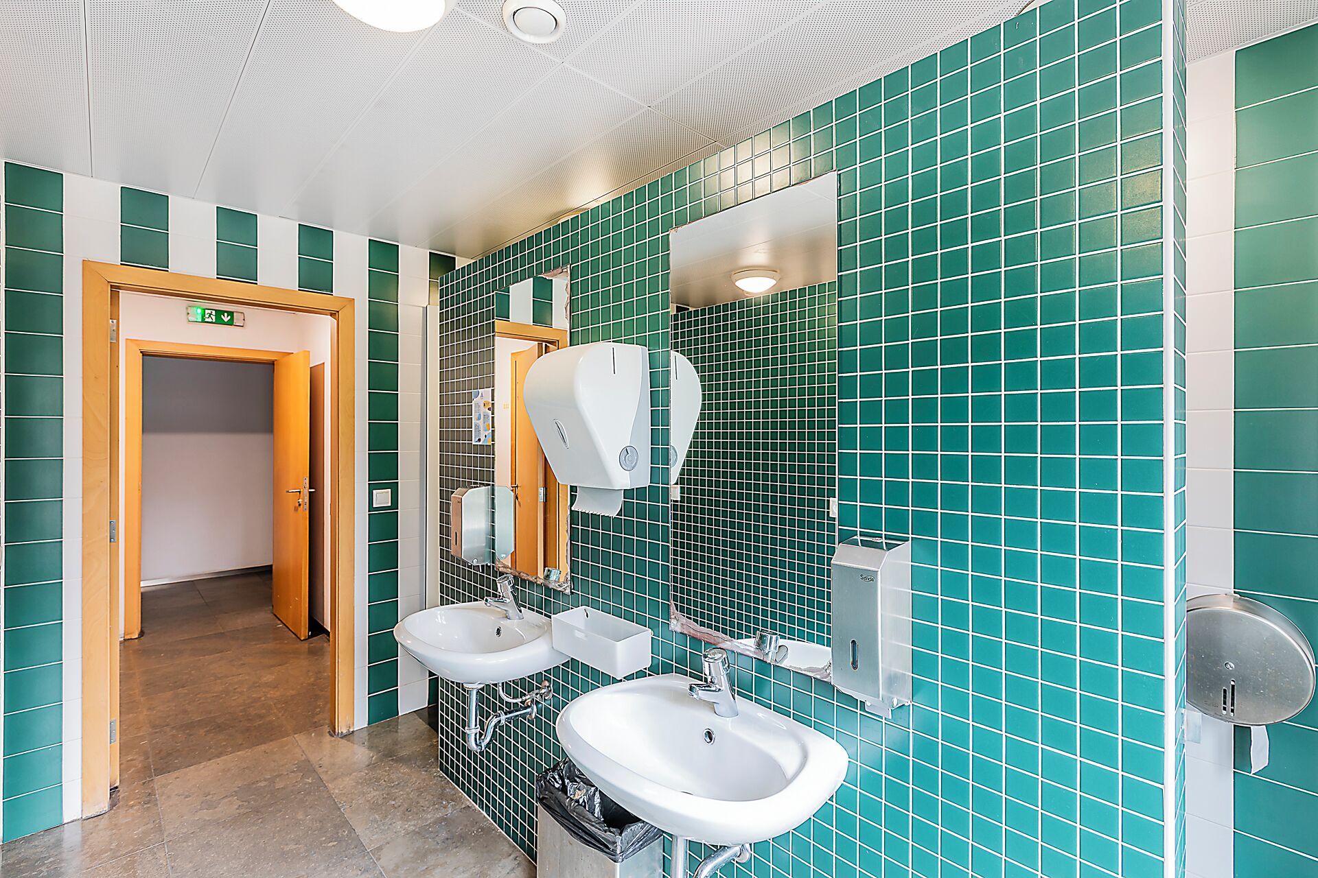 Un baño con paredes de azulejos verdes, dos lavamanos, un espejo y un secador de manos.