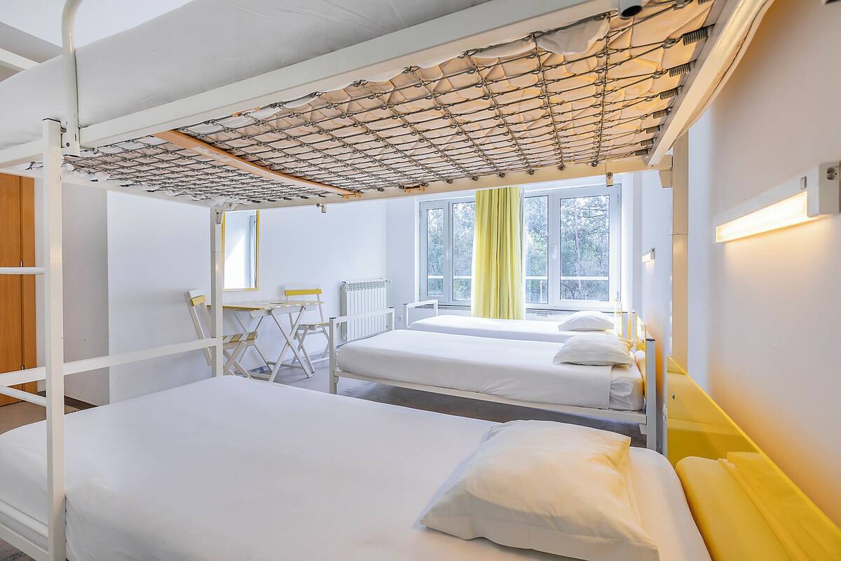 Ein Zimmer mit Etagenbetten, einem kleinen Tisch, Stühlen und einem Fenster mit Vorhängen.