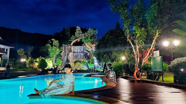 A nighttime pool area with a person sitting on the edge, surrounded by trees and a waterfall feature in the background. - Akantuka Homestay (Makham)