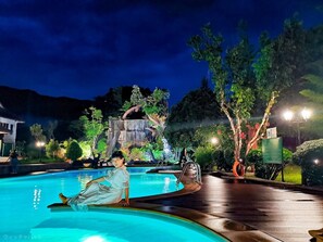 A nighttime pool area with a person sitting on the edge, surrounded by trees and a waterfall feature in the background. - Akantuka Homestay (Makham)