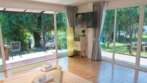 A bedroom with a large bed, a TV, and a view of a garden through large windows. - Akantuka Homestay (Makham)