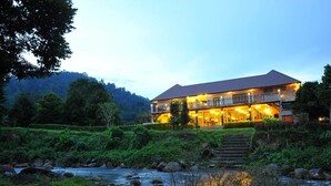 A two-story building with a balcony, surrounded by lush greenery and a river. - Akantuka Homestay (Makham)