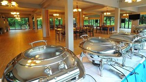 A buffet setup with multiple stainless steel chafing dishes on a table, surrounded by dining tables and chairs in a spacious room with large windows and chandeliers. - Akantuka Homestay (Makham)