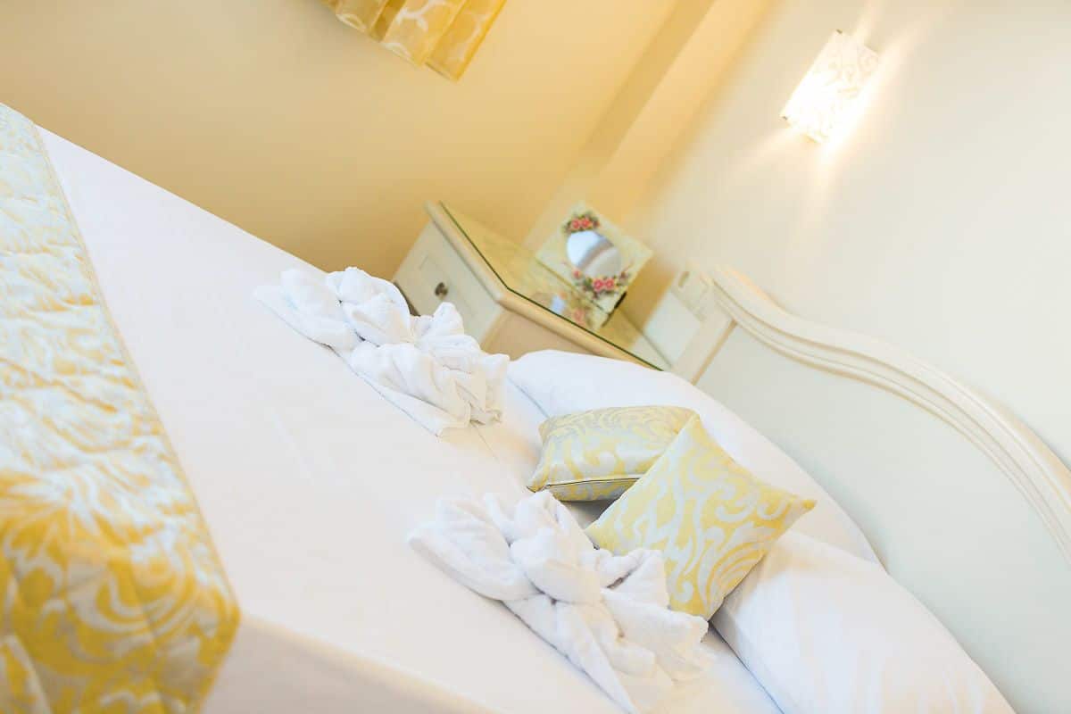A hotel room with a neatly made bed, a bedside table with a mirror, and a wall-mounted light fixture.