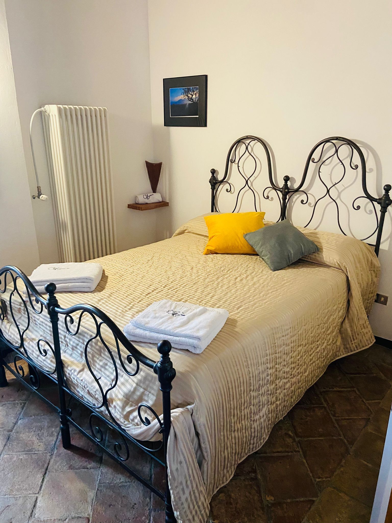 Cama bem arrumada com colcha listrada, um travesseiro amarelo e um travesseiro cinza. Uma toalha branca dobrada está sobre a cama. Uma moldura com um quadro está pendurada na parede acima da cama.