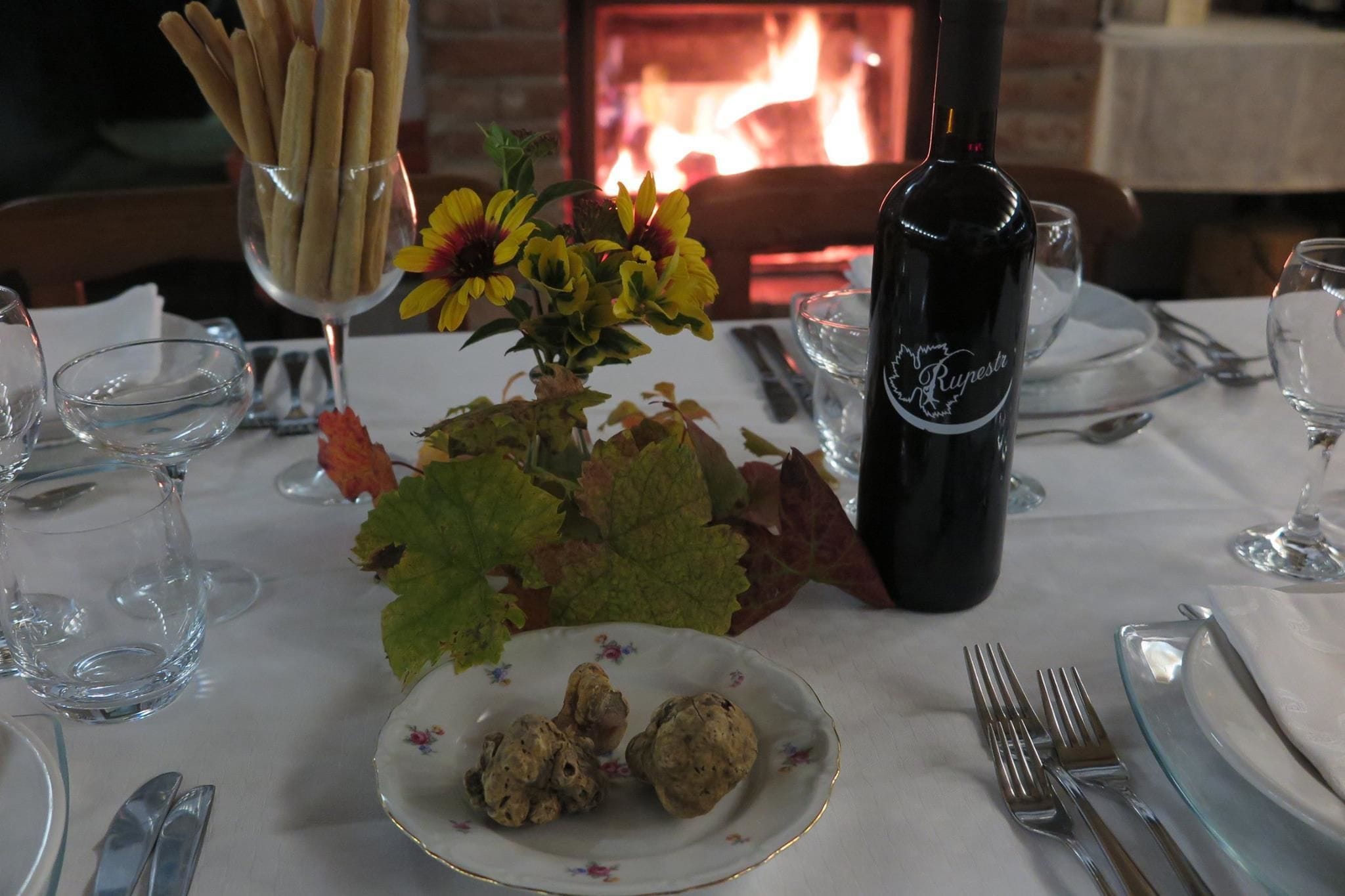 A dining table set with a bottle of wine, a plate of nuts, a glass vase with flowers, and a bowl of breadsticks.
