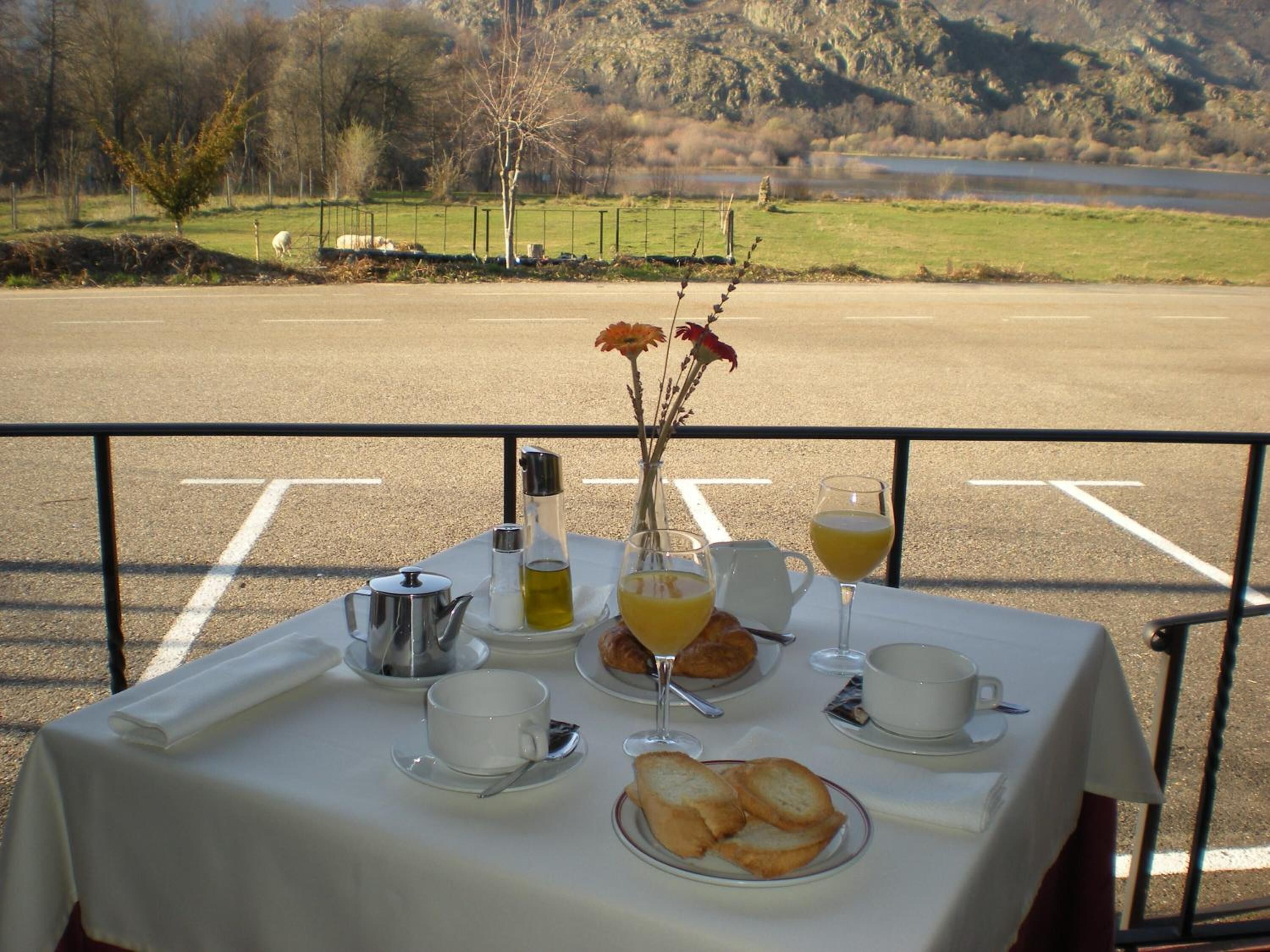 A table set with breakfast items including croissants, juice, and coffee, overlooking a scenic landscape with a lake and hills.