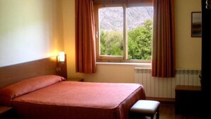 Una habitación con una cama, un banco, una ventana con cortinas y una lámpara de pared.