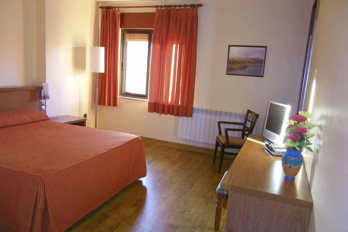 A hotel room with a bed, a desk with a computer and a vase of flowers, a chair, and a window with curtains.
