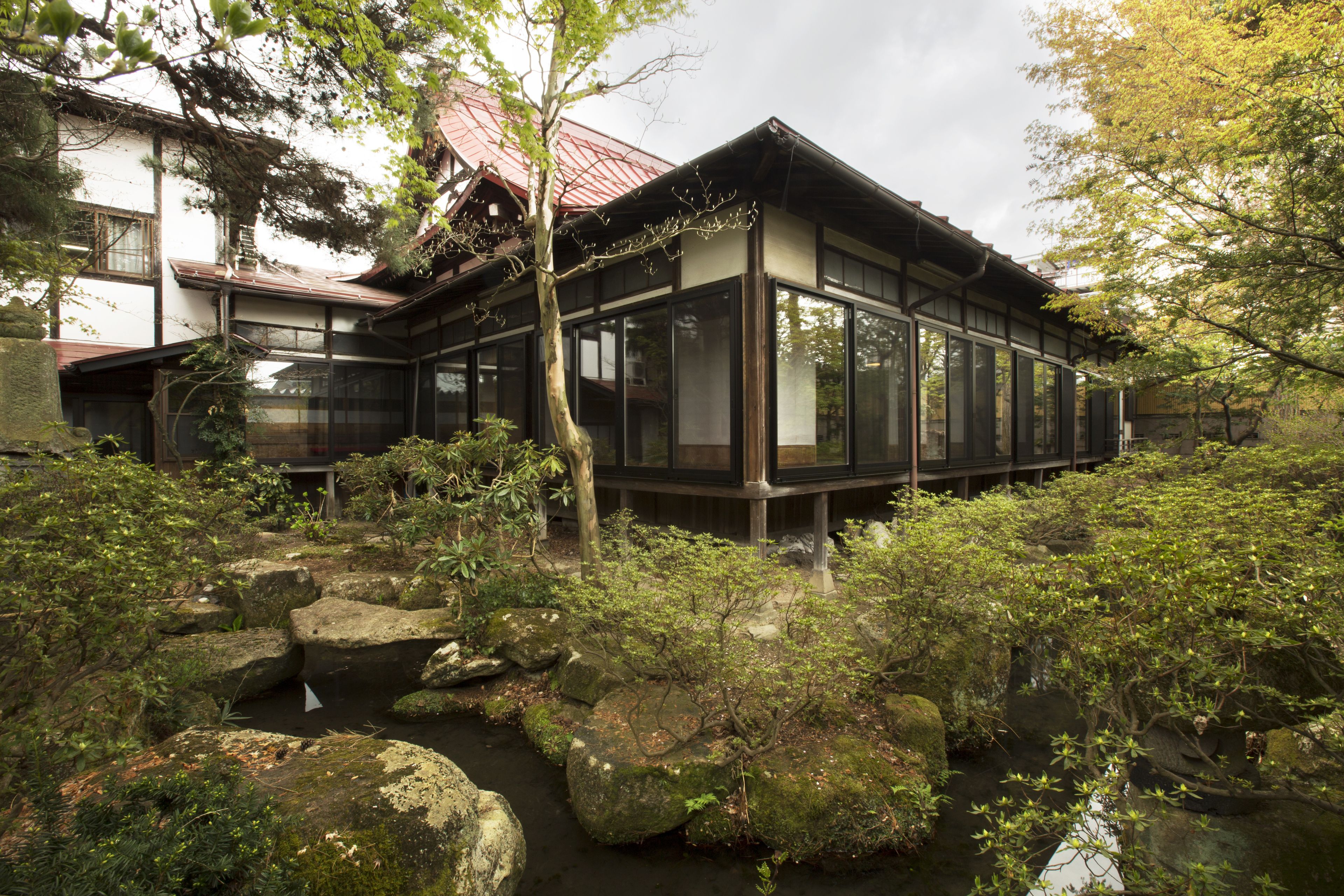 A traditional Japanese house with a wooden exterior and large glass windows, surrounded by a natural garden with rocks and trees.