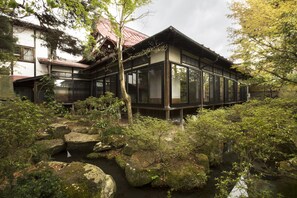 Une maison traditionnelle japonaise, avec une façade en bois et de grandes fenêtres en verre, entourée d’un jardin naturel agrémenté de rochers et d’arbres.