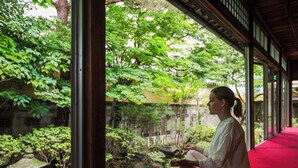 A person sitting on a red carpet in a room with large windows overlooking a garden with a stone lantern and greenery. - Temple Hotel Takayama Zenkoji (Takayama)