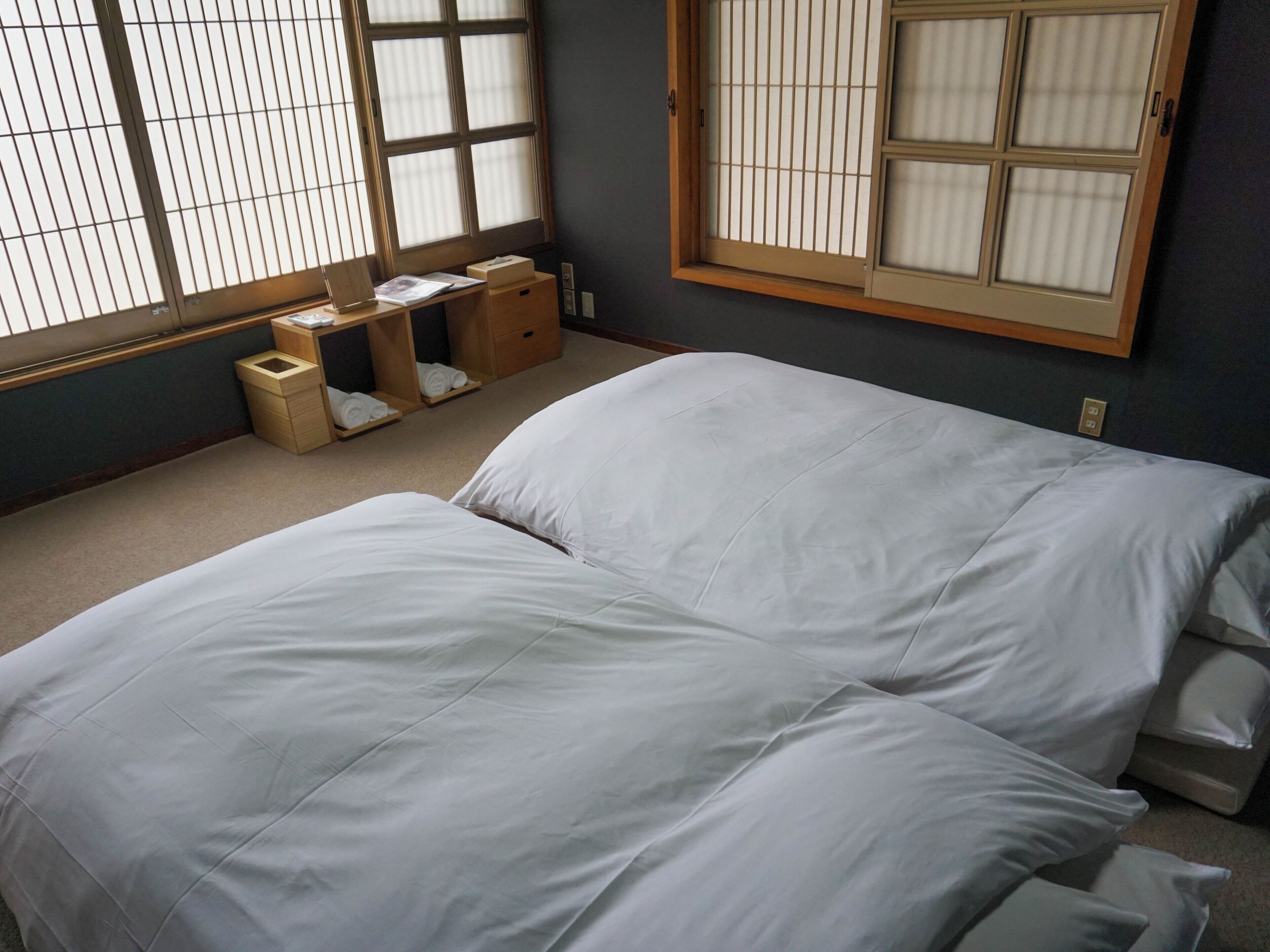 A hotel room with a bed, a desk with a chair, a window with blinds, and a small wooden cabinet.