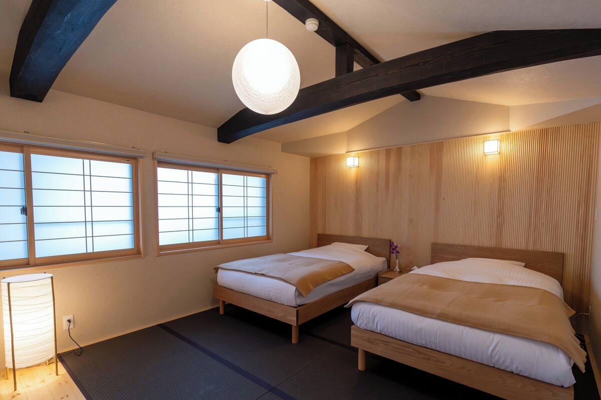 Habitación de hotel con dos camas de madera, suelo de esterilla de tatami, ventana con paneles de shoji y una lámpara en el techo.