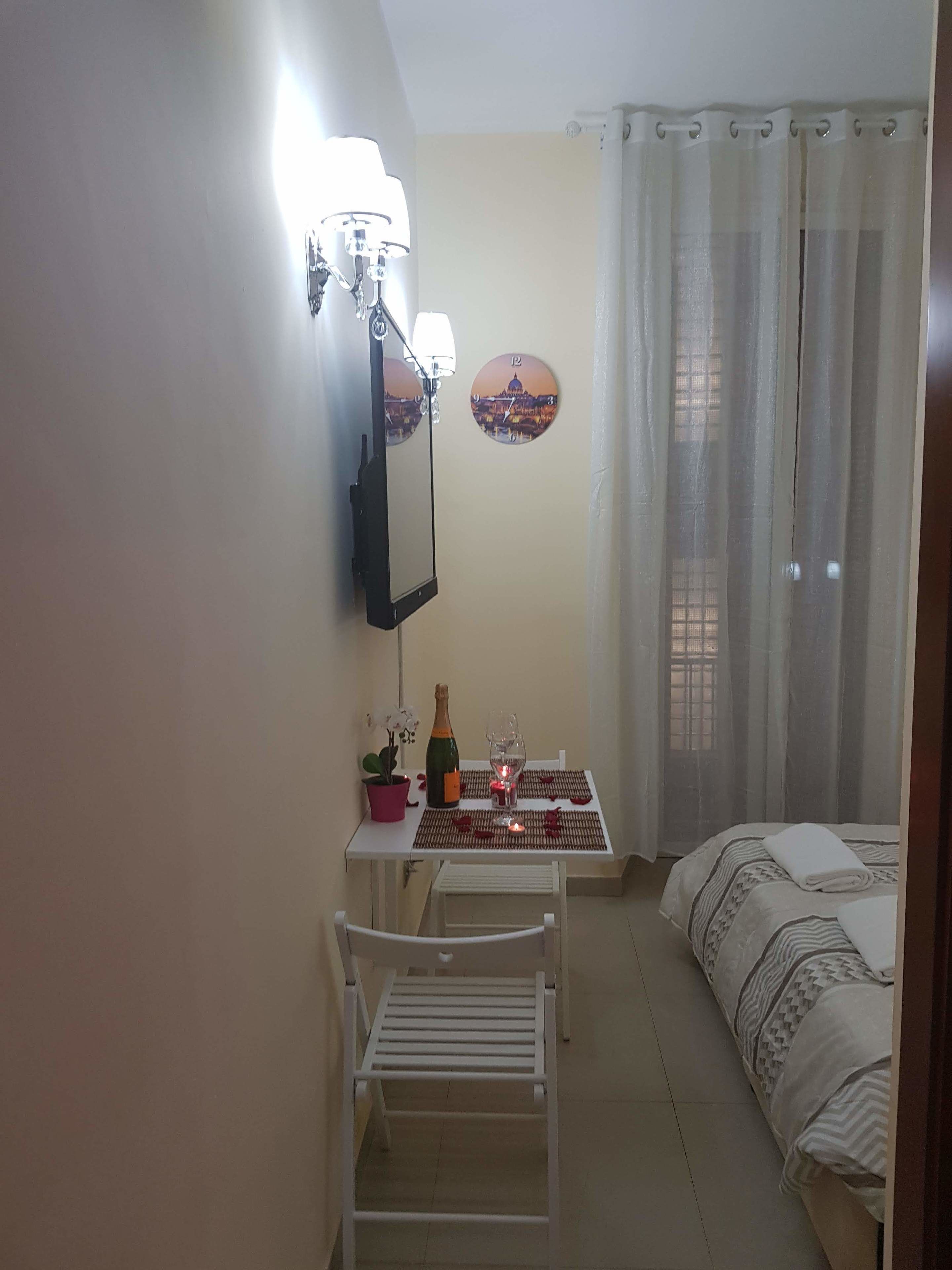 Una habitación pequeña y bien iluminada con una cama, una mesa con una botella y un vaso, una silla, un espejo y un reloj de pared.