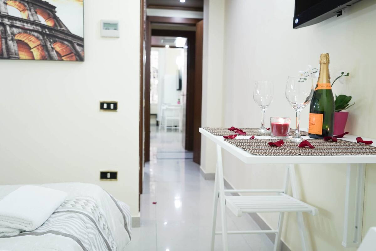 Habitación de hotel con una cama, una mesa dispuesta con una botella de champán, copas y una vela.