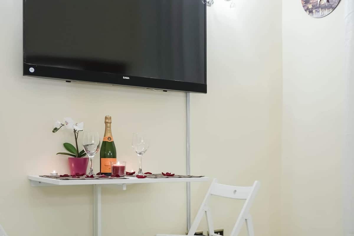 Habitación de hotel con una cama, un televisor montado en la pared, una pequeña mesa con una botella de champán, copas, una planta en maceta y una silla blanca.