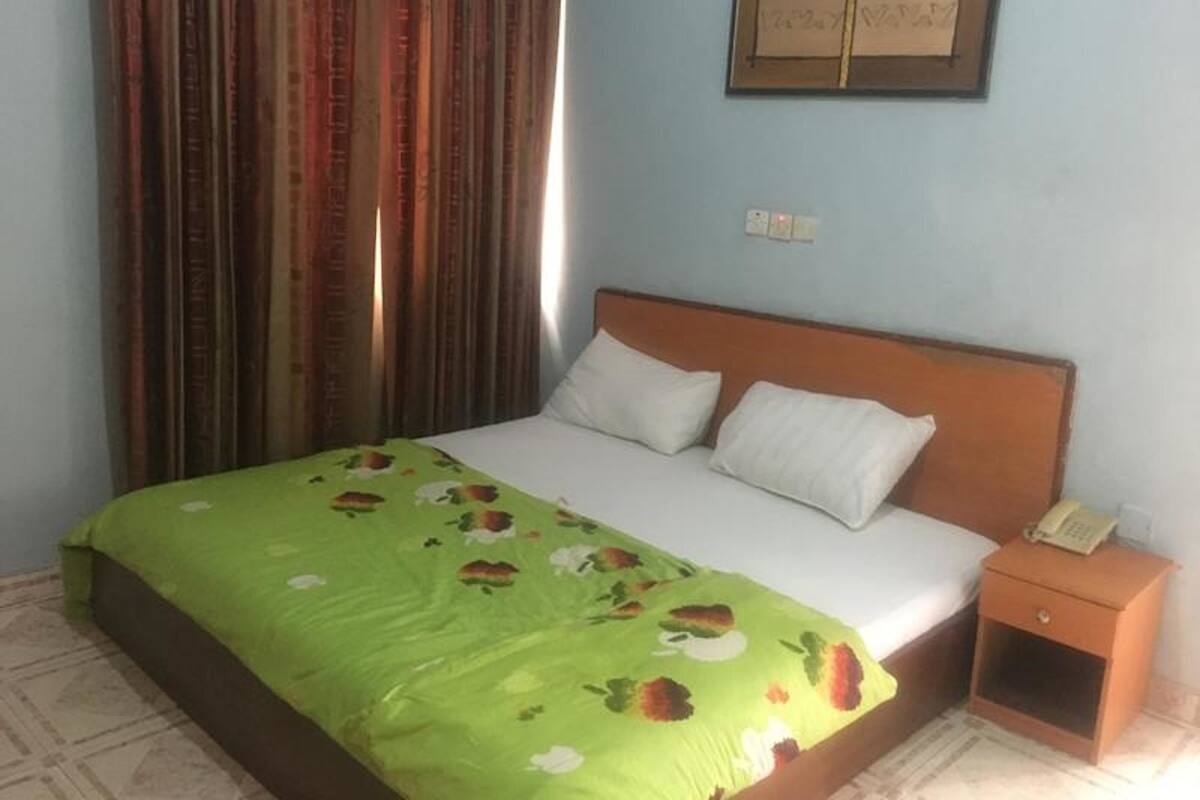 A room with a bed, a nightstand, a curtain, and a wall-mounted item.