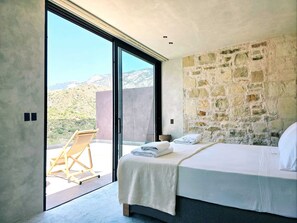 A bedroom with a large bed, a stone wall, and a view of a mountainous landscape through a sliding door. - Monastery Estate Mountain Retreat (Kantanos-Selino)