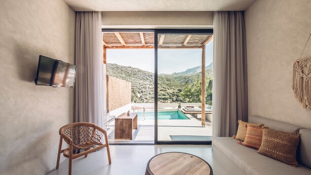 Une chambre moderne dotée d’une grande porte-fenêtre coulissante donnant sur un espace piscine, d’un canapé, d’un fauteuil et d’un téléviseur fixé au mur.