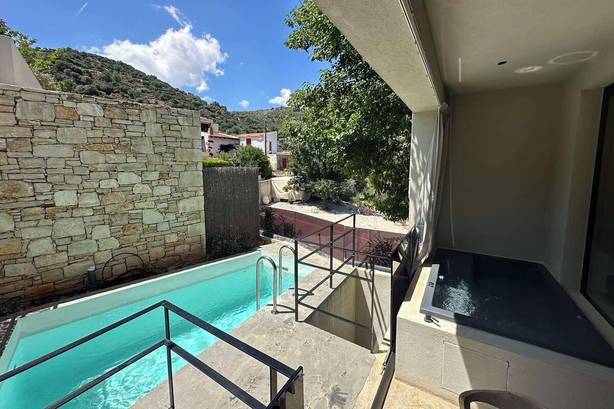 Zona de piscina con paredes de piedra, piscina azul cristalina y vistas de edificios y colinas circundantes.