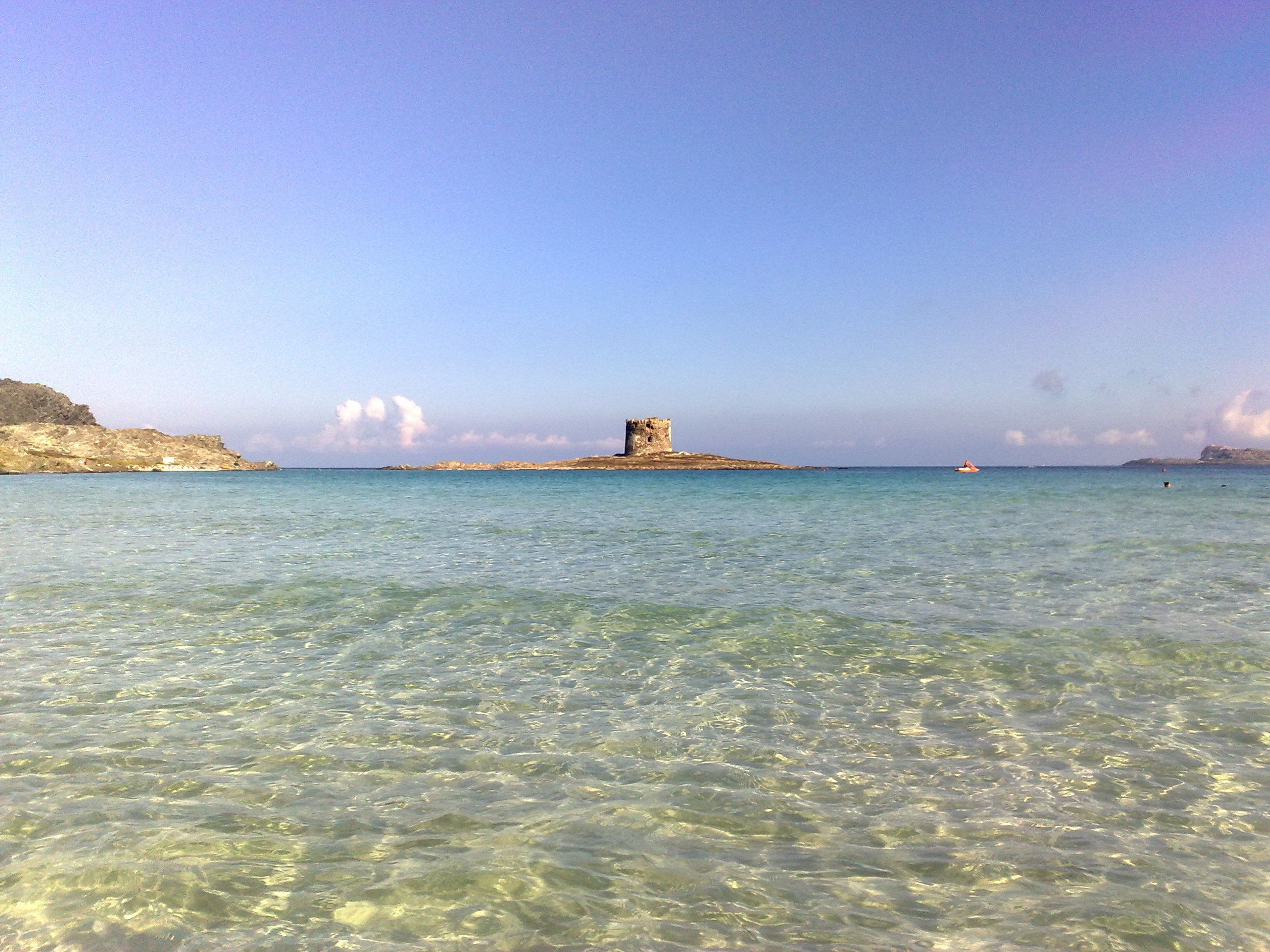 A coastal scene with clear shallow waters, a distant rocky outcrop, and a small island with a structure.