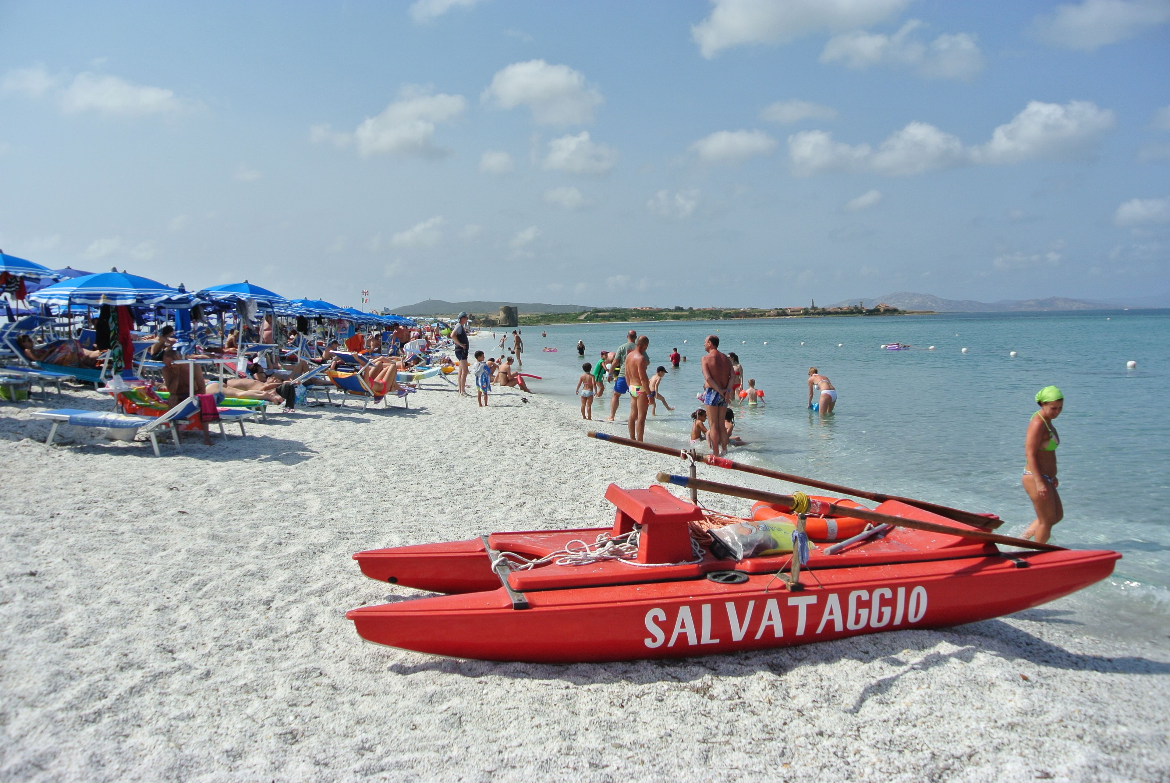 Ein Strand mit einem roten Boot, auf dem 'SALVATAGGIO' steht, und Menschen, die das Wasser genießen.